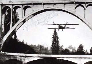 The black and white shot of a plane flying under a bridge